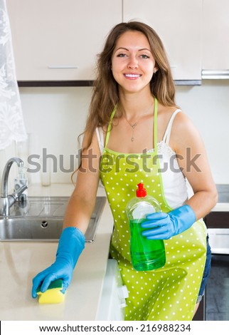Young beautiful smiling girl in apron cleaning furniture in kitchen