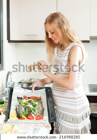 Smiling woman putting  saltwater fish into sheet pan at kitchen table