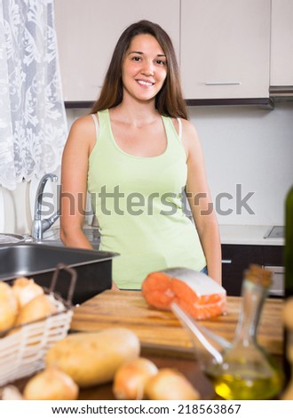 Young smiling woman cooking salmon fish in frying pan at kitchen