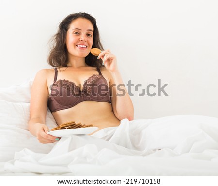 Smiling brunette girl eating chocolate chip cookies in her bed at home