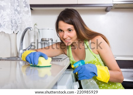 Happy young housewife in apron cleaning furniture in kitchen at home
