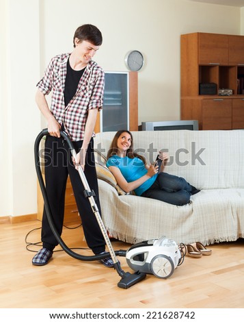 Young man cleaning with vacuum cleaner while wife lying with eBook over sofa at home