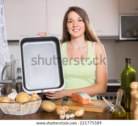 Smiling young woman cooking salmon fish in frying pan at kitchen