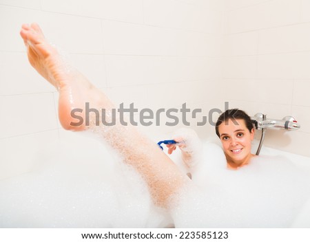 Smiling young brunette woman shaving legs in foam at bathtub