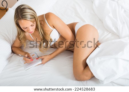 Blonde long-haired girl writing in notebook on white sheet in bed at home