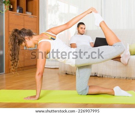 Young woman doing yoga at home and her boyfriend resting on couch with laptop