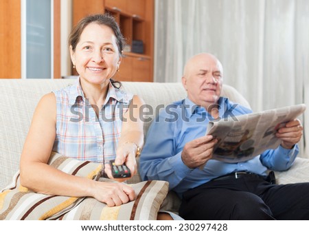 Portrait of  mature woman with TV remote against elderly man with newspaper in home interior