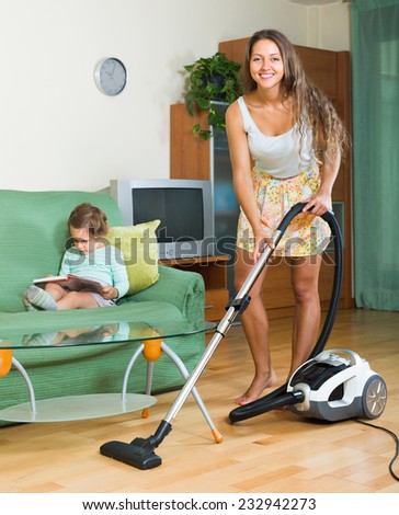 Smiling mother and little daughter chores with vacuum cleaner in home. Focus on woman