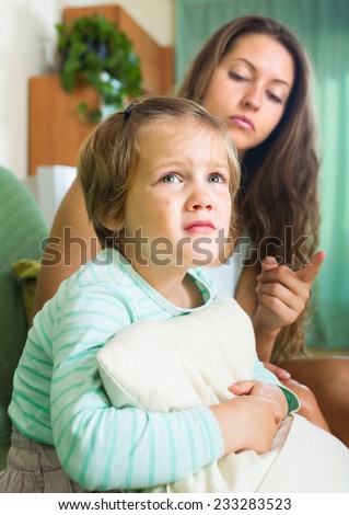 Unpleased young mother scolding little daughter in the living room at home. Focus on girl