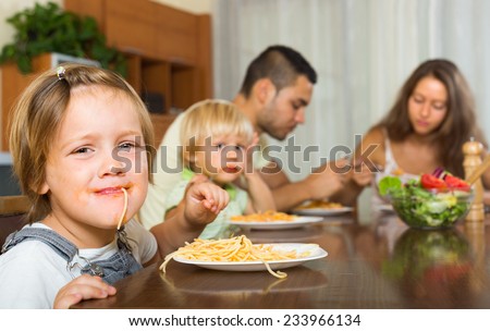 Smiling family with two little children eating with spaghetti at table. Focus on girl