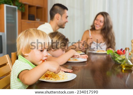 Happy smiling young family of four eating spaghetti at home interior. Focus on girl