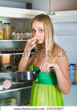 Girl holding her nose because of bad smell from food near refrigerator at home