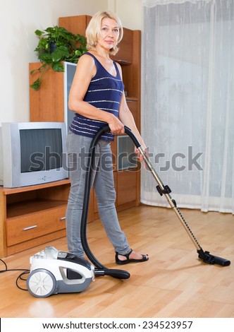 cheerful  mature woman cleaning with vacuum cleaner on parquet floor at home
