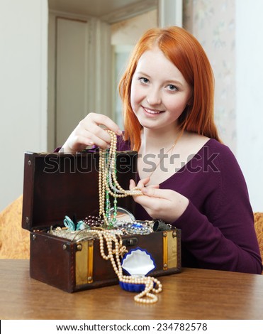 red-headed teenager girl looks jewelry in treasure chest at home interior