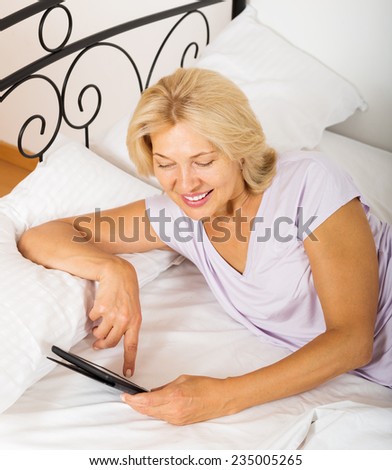 Cheerful  mature woman resting with ereader laying on bed at home