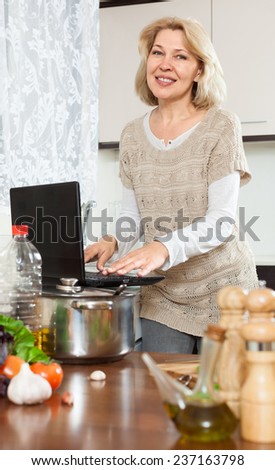 Elderly woman using laptop while cooking soup  at home kitchen