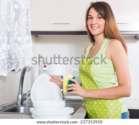 Happy smiling girl in apron washing plates with sponge in domestic kitchen