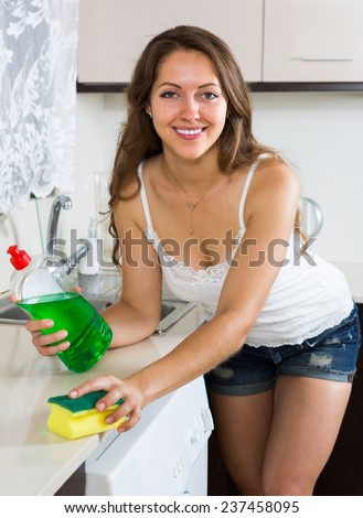 Smiling beautiful young housewife cleaning furniture in kitchen at home