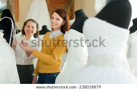 Shop assistant  helps to girl chooses white bridal outfit  at shop of wedding fashion. Focus on young