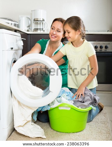 Home family laundry. Happy mother with preschooler daughter loading clothes into washing machine in kitchen. Focus on girl