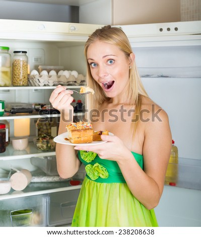 Happy hungry woman near opening fridge eating cake in her home
