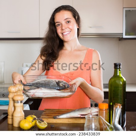 Smiling young brunette woman cooking fish in home kitchen