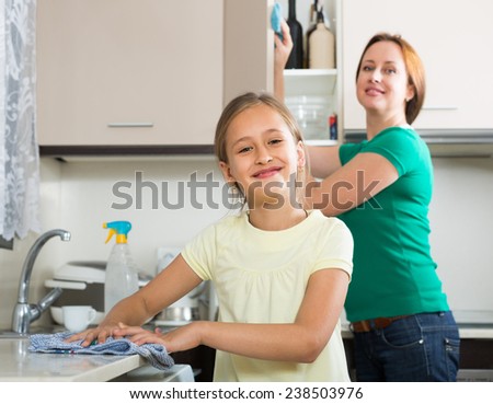 Happy schoolgirl helping mother dusting furniture indoor. Focus on girl