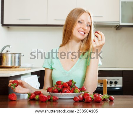 Positive blonde girl eating strawberry in home kitchen