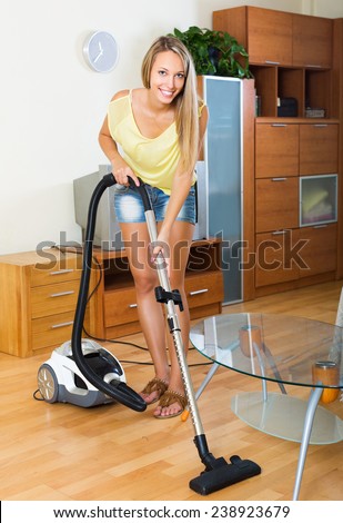 Blonde  woman in shorts cleaning with vacuum cleaner on parquet floor at home