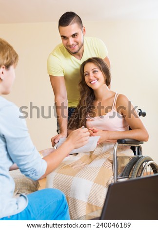 Female agent consulting smiling young woman in invalid chair and husband. Focus on woman