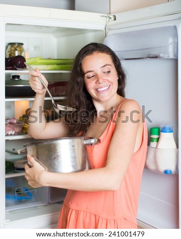 Hungry young brunette woman eating soup from pan near fridge at home