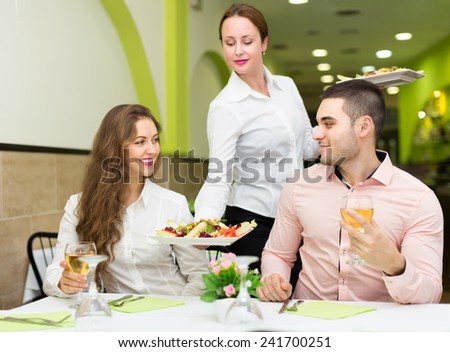 Smiling female waiter with plates serving guests table in cafe. Focus on girl