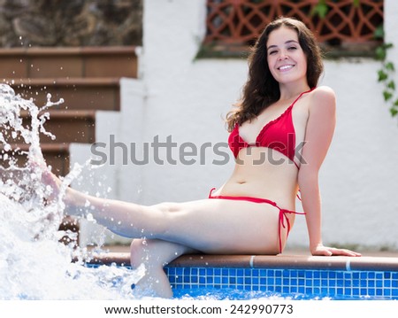 Happy brunette woman in red bikini making splash with water in pool