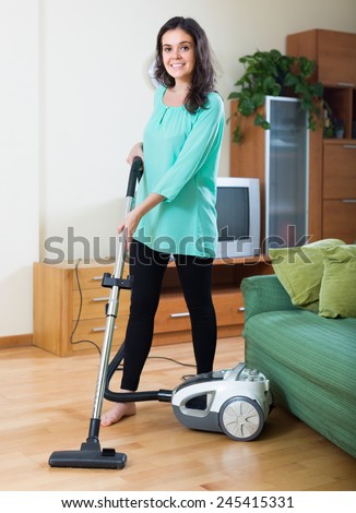 Smiling young brunette woman cleaning living room with vacuum cleaner