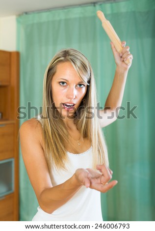 Angry young housewife with rolling-pin in hand complaining at home