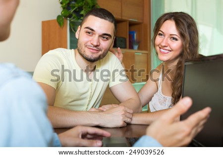 Smiling young married couple talking with employee with laptop at home