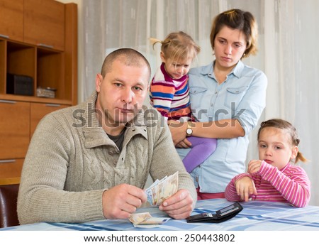Family arguing about money inside the house. Depressed husband is sitting on a sofa and his wife with their child is standing behind him