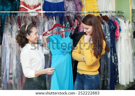 pretty girl chooses evening dress at clothing boutique. Friendly shop consultant helps her