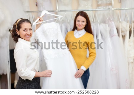 Woman helps the bride in choosing bridal gown at shop of wedding fashion. Focus on mature