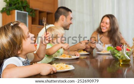 Positive young family of four eating with spaghetti at table. Focus on girl