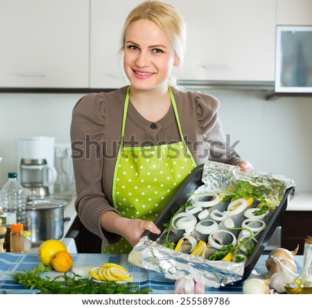 Happy smiling blonde woman cooking fish with lemon in kitchen at home