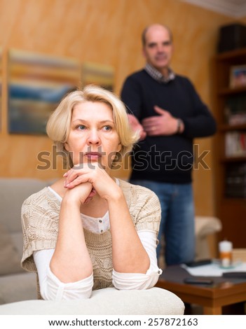 Pensioners couple are having a quarrel. Mature wife is sitting turned away from her old husband with her arms near her cheek and the husband is standing behind and talking to her