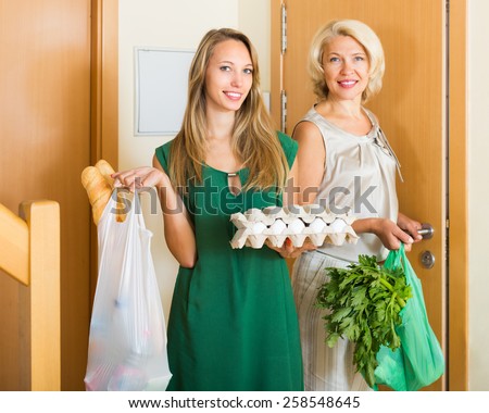 Mature mother and adult daughter holding bags of food near apartment. Focus on young