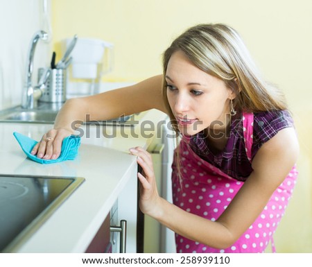 Smiling blonde girl in apron cleaning furniture in kitchen