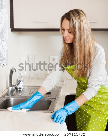 Blonde woman in apron cleaning  furniture with rag in kitchen at home