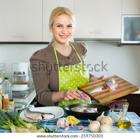 Happy smiling woman putting pieces of fish in frying pan at home kitchen