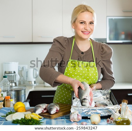 Smiling blonde woman putting pieces of lemon in fish at home kitchen