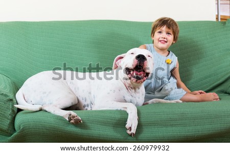 Happy smiling little girl with friendly big white dog on sofa at home. Focus on dog