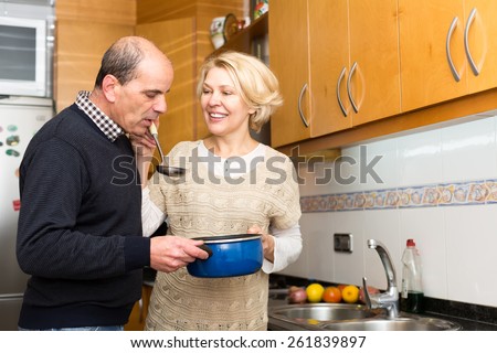Mature family couple cooking dinner in kitchen. Senior woman gives her man soup in a ladle to taste