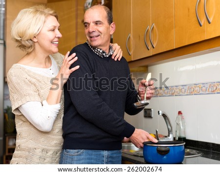 Happy senior man cooking soup for his beautiful elderly wife in kitchen. His wife hugs him from his back